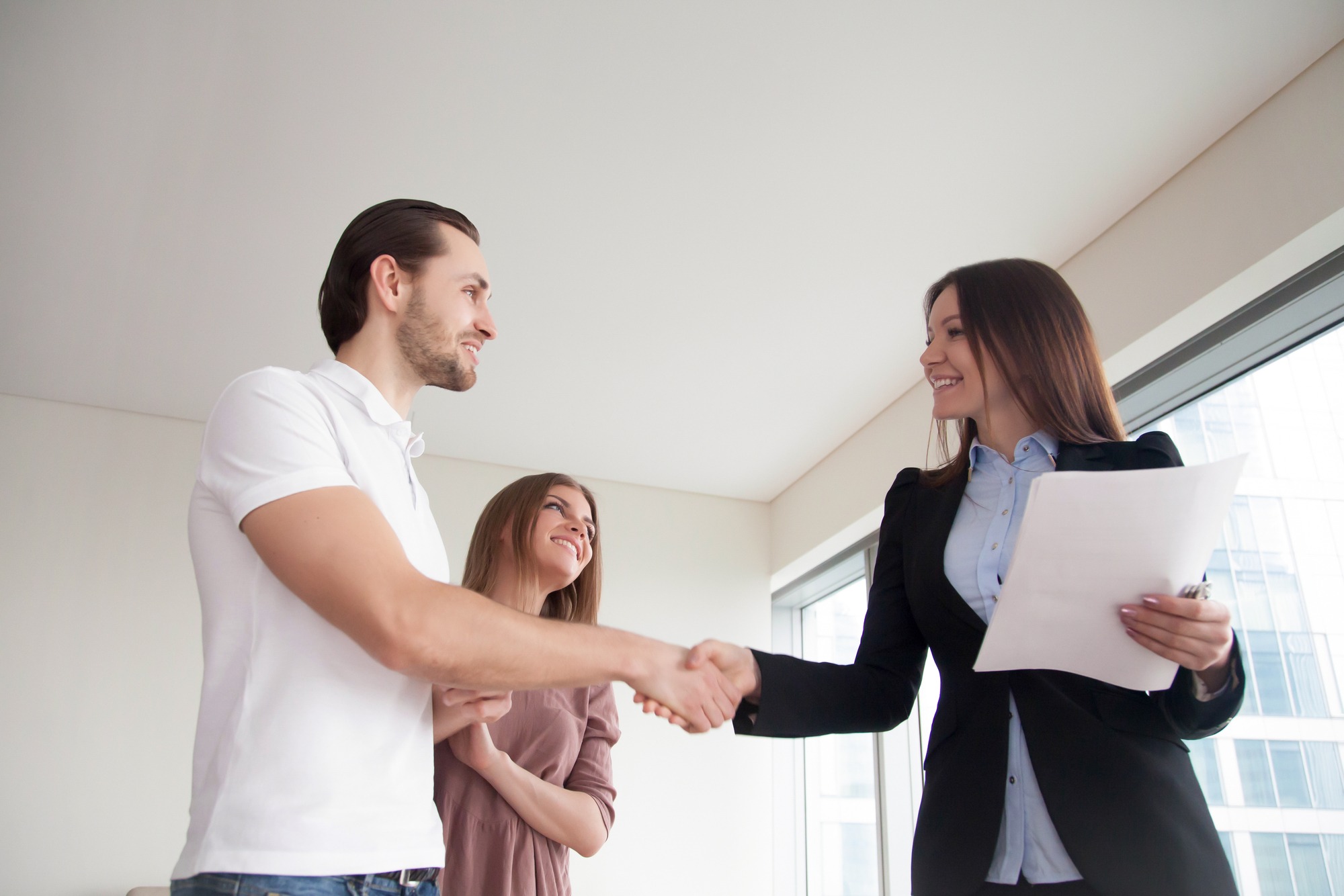 Real estate agent showing apartment to young man and woman, cheerful smiling couple shaking hands with friendly realtor holding papers